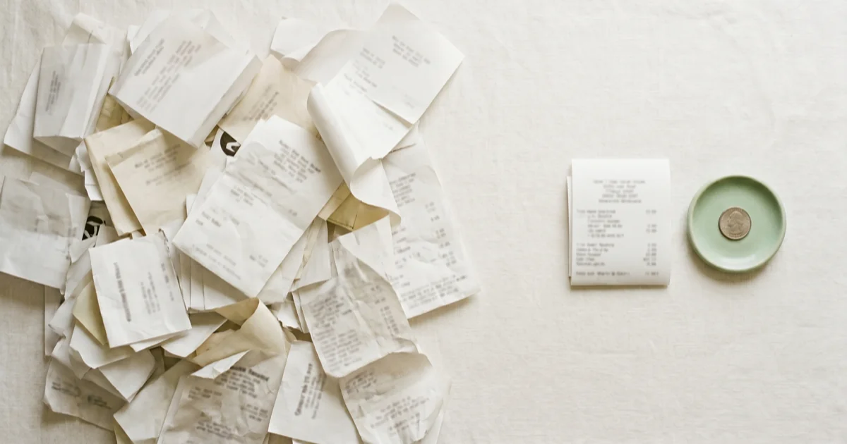 Flat-lay of a tangled pile of paper receipts beside a clean single receipt and a small sage-green ceramic dish holding a coin